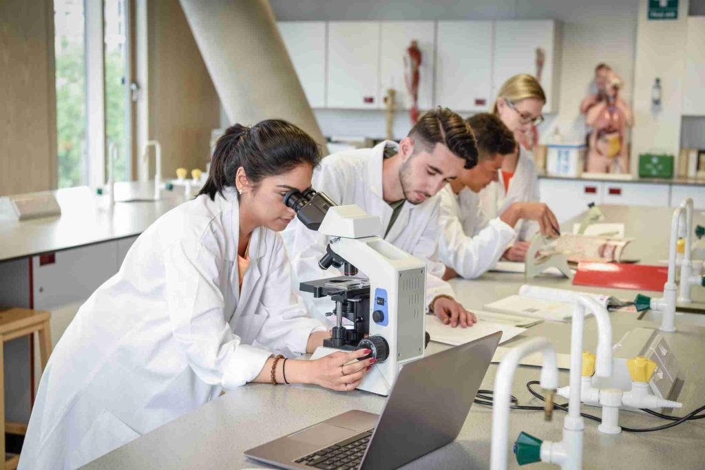 imagem de um laboratório onde estão profissionais de jaleco branco debruçados em uma mesa vendo microscópio, papeis, peças anatômicas e outros.