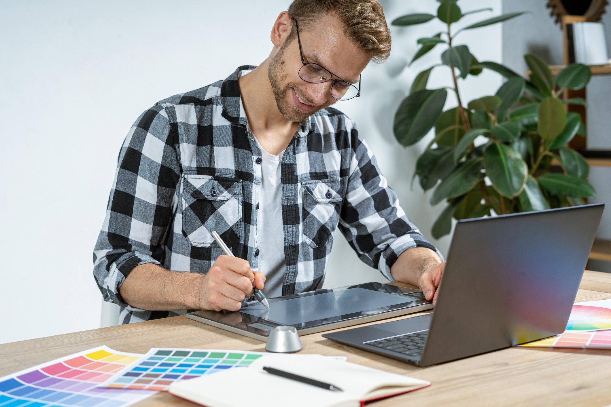 homem branco, loiro, de camisa xadrez preta está sentado na frente de uma mesa que tem um notebook, papeis coloridos e um tablet no qual ele usa para desenhar com uma caneta especial