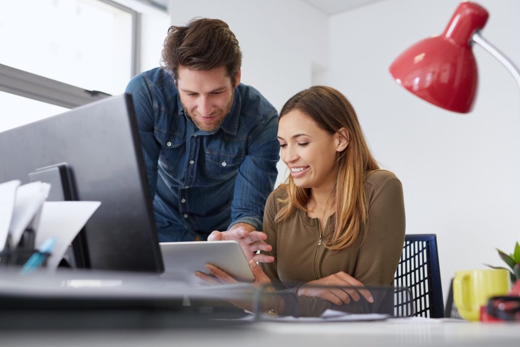 mulher está sentada em uma mesa de escritório segurando um tablet e olhando pra ele; ao lado está de pé curvado na mesa um homem com a mão no mesmo tablet.