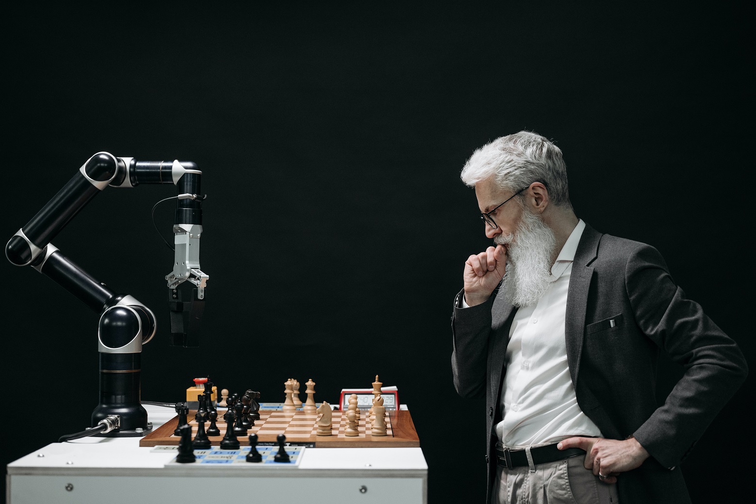 um homem e um robô jogam xadrez. Em uma mesa branca está o tabuleiro aberto com as peças e o robô está jogando enquanto o homem o observa